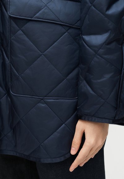 Τσέπη και μανίκι σκούρου μπλε μπουφάν με ραφές, με το χέρι να ακουμπά κοντά στο τελείωμα πάνω σε απλό φόντο.