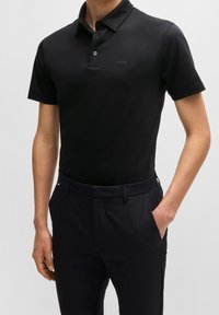 Homme portant un polo noir ajusté à manches courtes avec le logo "BOSS" sur la poitrine, associé à un pantalon noir, les mains dans les poches.