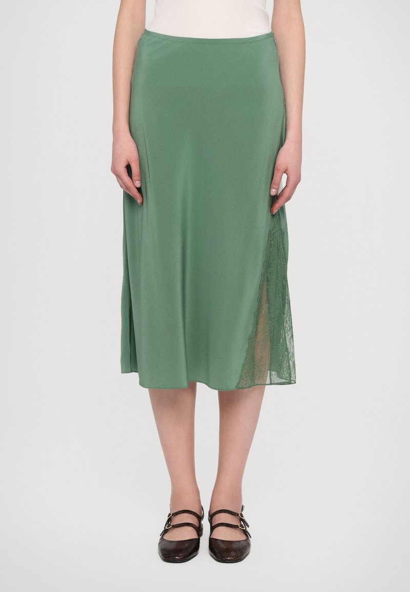 Groene satijnen midi rok met kantpaneel detail aan de zijkant. Gladde textuur, A-lijn vorm en een schone afwerking. Draagt met donkerbruine schoenen.