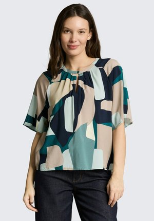 Kobieta o długich brązowych włosach, ubrana w luźną, krótką bluzkę z abstrakcyjnymi geometrycznymi wzorami w kolorach morskim, granatowym, beżowym i czarnym, w połączeniu z ciemnymi dżinsami.