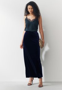 Camisole in raso nero con bordatura in pizzo, abbinata a una gonna lunga in velluto blu navy e tacchi neri con cinturino, che tiene una pochette con motivo dorato.
