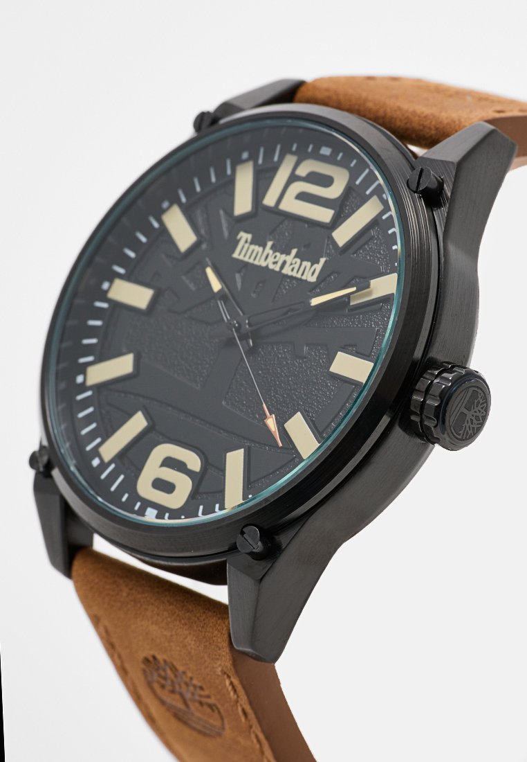 Reloj de pulsera negro con una esfera negra texturizada, grandes números y marcadores de horas, y correa de cuero marrón. Presenta una corona con la marca y acentos.