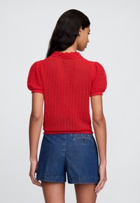 Sarkans adīts tops ar pūkainām piedurknēm un viļņotu izgriezumu, ar fakturētu rakstu. Kombinēts ar zilām džinsa �šortām.