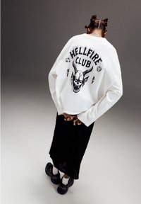 Bílá mikina s nápisem "HELLFIRE CLUB" a grafikou lebky na zádech, spárovaná se černou sukní a černými teniskami.