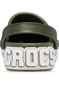 Zoccoli verdi in gomma Crocs con una cinghia testurizzata, caratterizzati da un logo "CROCS" spesso in bianco e grigio sul tallone. Design flessibile e leggero.