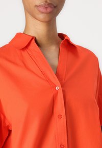 Camicia di cotone arancione brillante con chiusura a bottoni, colletto a V, maniche corte e una texture liscia. Dettaglio dei bottoni ben visibile sul collo.