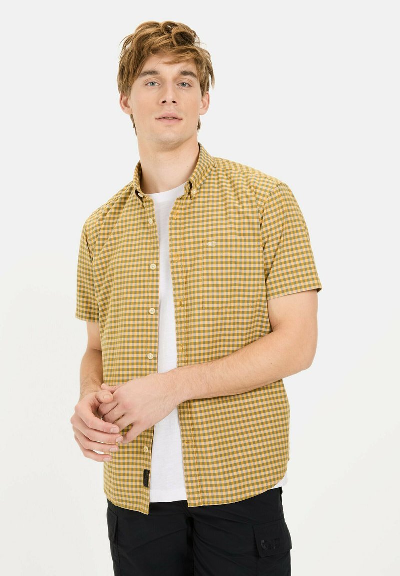 Korte mouwen, knoopshirt in een geel en bruin ruitpatroon, met een borstzak en een wit T-shirt eronder, gedragen met donkere broek.