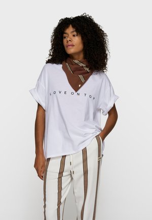 Kobieta o kręconych włosach, ubrana w białą koszulkę z podwiniętymi rękawami z napisem "LOVE ON TOP", brązową paskowaną chustę na szyi oraz białe spodnie z brązowymi pionowymi paskami.