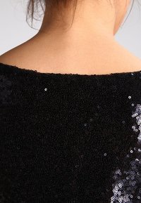 Top noir en sequins avec une surface texturée, dotée d'un décolleté ouvert et d'un dégradé de sequins brillants sur un côté.
