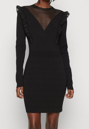 Nainen pukeutuneena istuvaan mustaan mekkoon, jossa on pitkät hihat, läpinäkyvä V-kaula-aukko ja röyhelödetaileja olkapäillä ja rinnalla.