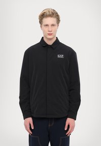 Svart lättviktsjacka med krage, knappstängning och EA7-logotyptryck på vänster bröst. Slät yta och avslappnad passform.
