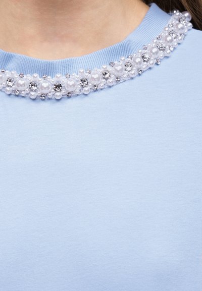 Κοντινό πλάνο σε λαιμόκοψη από ανοιχτό μπλε ύφασμα, διακοσμημένη με εναλλασσόμενες μεγάλες και μικρές λευκές πέρλες και διάφανα ρινίσματα.