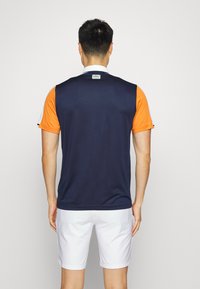 Mörkblå och orange golftröja med kort ärm och vit krage, med en logotags på baksidan och ett mjukt, lätt tyg.