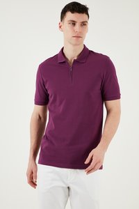 Muž oblečený v krátkemu rukávu fialovej polokošeli so zipsom a bielych nohaviciach, stojaci pred jednoliatym svetlým pozadím.