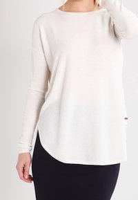 Blusa blanca de manga larga con tejido texturizado, escote redondo y un corte holgado. Presenta un dobladillo curvado y una pequeña etiqueta decorativa en el lateral.