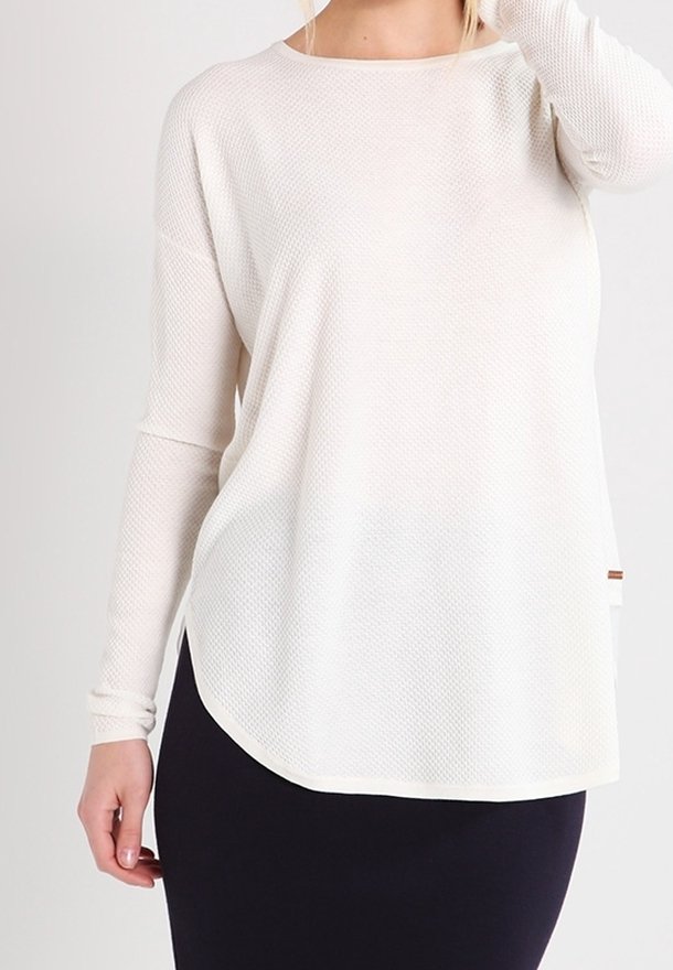 Blusa blanca de manga larga con tejido texturizado, escote redondo y un corte holgado. Presenta un dobladillo curvado y una pequeña etiqueta decorativa en el lateral.