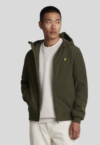 Olivengrønn zip-up jakke med hette, med en liten gul brodert logo på brystet, iført over en lys grå genser.