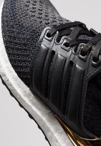 Černá pletená sportovní obuv s texturovaným svrškem, plastovými doplňky pro podporu, bílou tlumící mezipodešví a zlatým akcentem na boku.