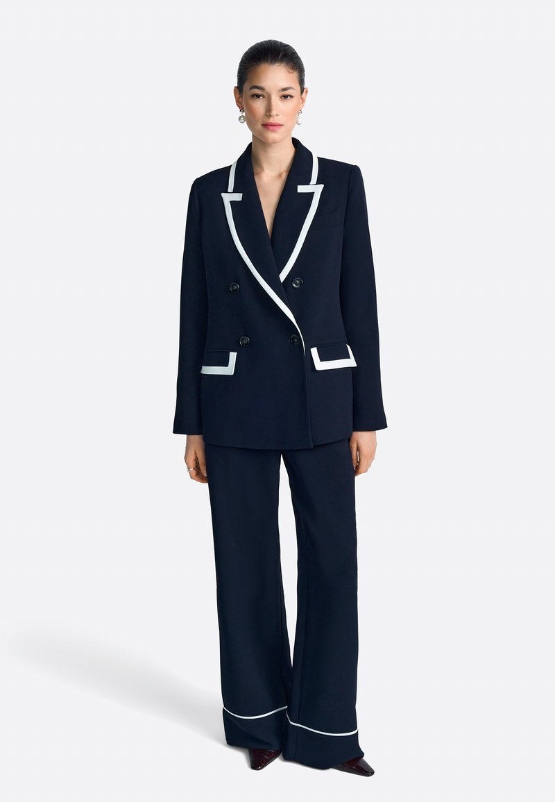 Blazer azul marino de doble botonadura con ribetes blancos, combinado con pantalones de pierna ancha azul marino con detalles blancos y elegantes bolsillos.