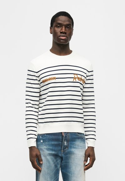 Homme portant un pull à manches longues blanc avec des rayures bleu marine, portant un texte orange, et un jean bleu délavé, debout devant un fond uni.