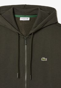 Sweat à capuche zippé vert foncé avec cordons, petit logo crocodile vert brodé sur la poitrine gauche, et étiquette "Lacoste Classic Fit" à l'intérieur du col.