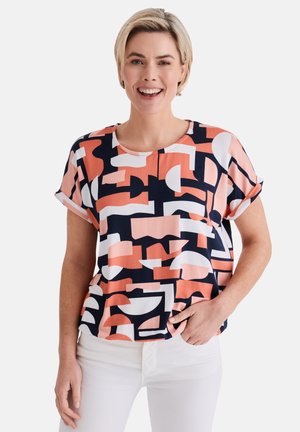 Smiling woman wearing a short-sleeve blouse with abstract coral, navy, and white geometric patterns, and white pants, standing against a plain background.