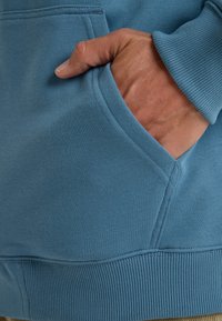 Sweatshirt bleu clair avec une texture douce, comportant une poche avant et des poignets côtelés. Les coutures sont nettes sans qu'il y ait de matériel visible.