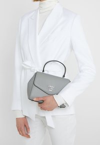 Szara skórzana torebka o ustrukturyzowanym kształcie, z połyskującą czarną rączką i klapą zamykającą. Na przedniej części znajduje się srebrne logo „DKNY”.