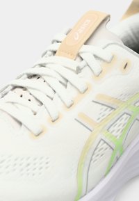 Zapato deportivo blanco con parte superior de malla, cordones planos y suela de goma. Cuenta con detalles en verde claro y gris. Fabricado con materiales sintéticos.