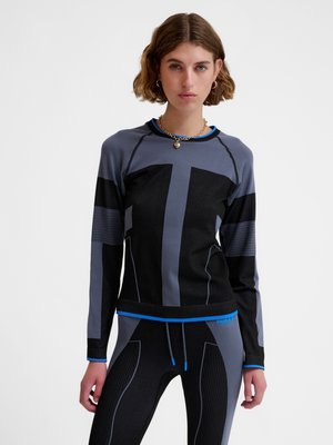 Vrouw met kort bruin haar, die een aansluitende zwarte en grijze sporttop en bijpassende leggings met blauwe rand draagt tegen een effen achtergrond.