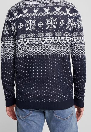Pull bleu marine avec des flocons de neige blancs, des rennes et des motifs géométriques porté par une personne en jean bleu clair, vu de dos.