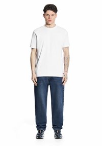 Biała koszulka z krótkim rękawem w zestawieniu z ciemnoniebieskimi dżinsami i czarnymi, wzorzystymi butami sportowymi. Model stoi, ręce wzdłuż ciała.