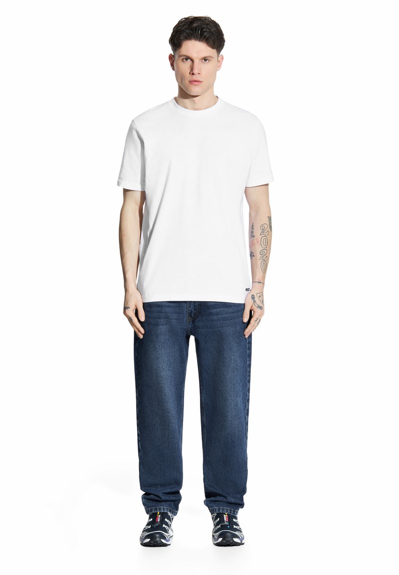 Biała koszulka z krótkim rękawem w zestawieniu z ciemnoniebieskimi dżinsami i czarnymi, wzorzystymi butami sportowymi. Model stoi, ręce wzdłuż ciała.