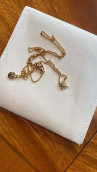 Colier cu lanț de culoare aurie cu pandantiv în formă de stea, având o piatră prețioasă clară, așezat pe un săculeț alb texturat.