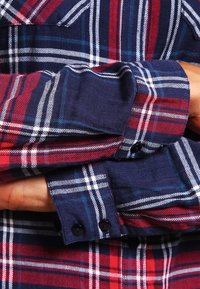 Dettaglio della camicia a quadri rossi e blu navy che mostra i polsini con bottoni, il motivo a scacchi contrastante e il tessuto strutturato con strisce blu e bianche.