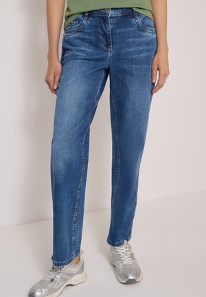 Person trägt blaue Jeans mit geradem Bein und silberne Turnschuhe und steht vor einem neutralen Hintergrund.