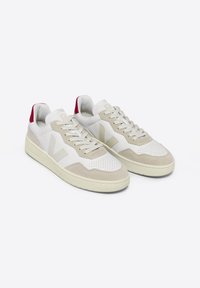 Baskets blanches et beiges avec un design perforé, des accents en suède doux et un détail rouge sur le talon. Elles sont équipées d'un système de laçage et de semelles en caoutchouc texturé.