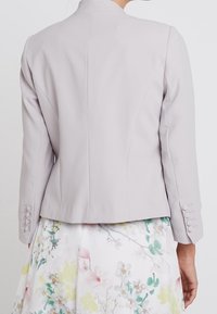 Blazer gris clair à coupe structurée, avec col montant, manches longues et détails de boutons aux poignets, assorti à une jupe fleurie.