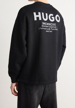 Persoon die een zwarte trui draagt met grote witte "HUGO" tekst en kleinere handelsmerk- en oprichtingsgegevens op de bovenrug.