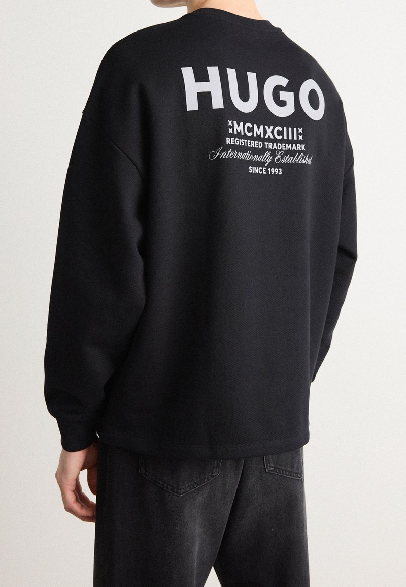 Personne portant un sweat-shirt noir avec un grand texte blanc "HUGO" et des détails plus petits de marque déposée et d'établissement sur le haut du dos.