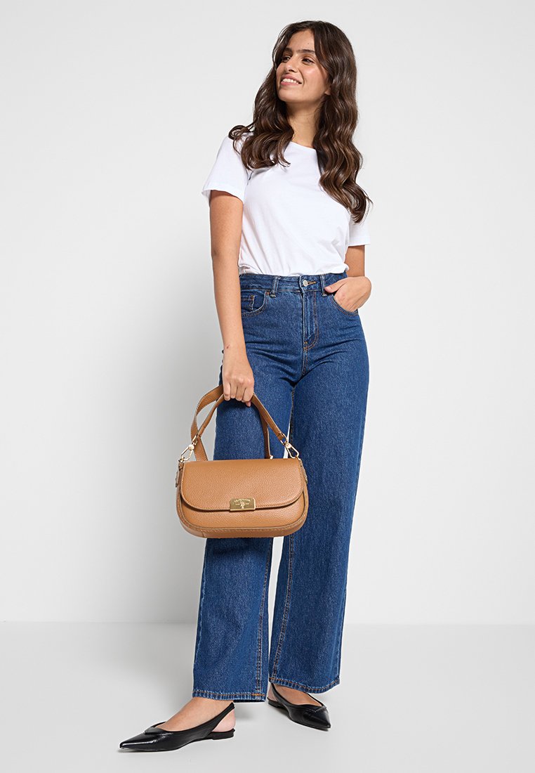Sac en cuir marron avec une texture lisse, un rabat sur le dessus et des accessoires en or, tenu par une personne portant une chemise blanche et un jean taille haute bleu.