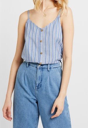 Kobieta ubrana w niebiesko-biały, pionowo paskowany bezrękawnik z guzikami, wpuszczony w luźne, niebieskie, wysokiej talii dżinsy.