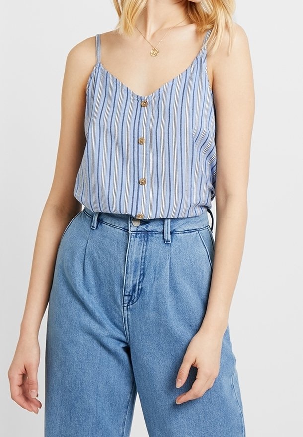 Femme portant un haut sans manches boutonné à rayures verticales bleues et blanches, rentré dans un jean bleu ample taille haute.