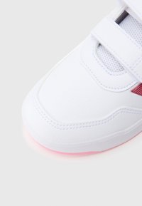 Biały but sportowy z siateczkowymi wstawkami, paskami na rzep i teksturowanym wykończeniem. Podeszwa ma delikatny różowy akcent.