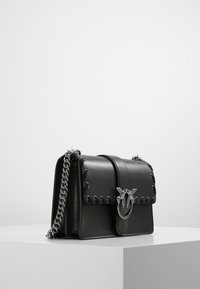 Černá kožená kabelka se stříbrným řetízkovým popruhem, dekorativní stříbrnou sponou ve tvaru ptáka a pletenými koženými detaily na klopě, umístěná na bílé ploše.