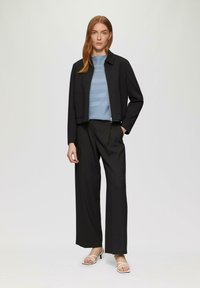 Schwarze cropped Jacke mit einem Reißverschluss vorne, kombiniert mit einem hellblauen gerippten Pullover und schwarzen weit geschnittenen Hosen. Weiße Riemchen-Heels.