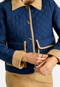 Granatowa pikowana kurtka z beżowym kołnierzem z sztruksu, z dwoma przednimi kieszeniami, zapięciem na guziki i kontrastowymi wykończeniami.