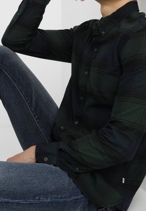 Person siddende med det ene knæ op, iført en mørkegrøn og sort ternet skjorte med knapper og blå denimjeans.