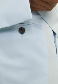 Svijetloplava tkanina s glatkom teksturom, s crnim zatvaranjem na četiri gumba i bočnom džepu sa suptilnim detaljima šivanja.