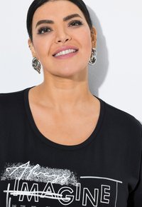 T-shirt nera con scollatura rotonda, caratterizzata da testo bianco e design grafico. Indossata con orecchini a cerchio argentati. Tessuto dalla consistenza liscia.